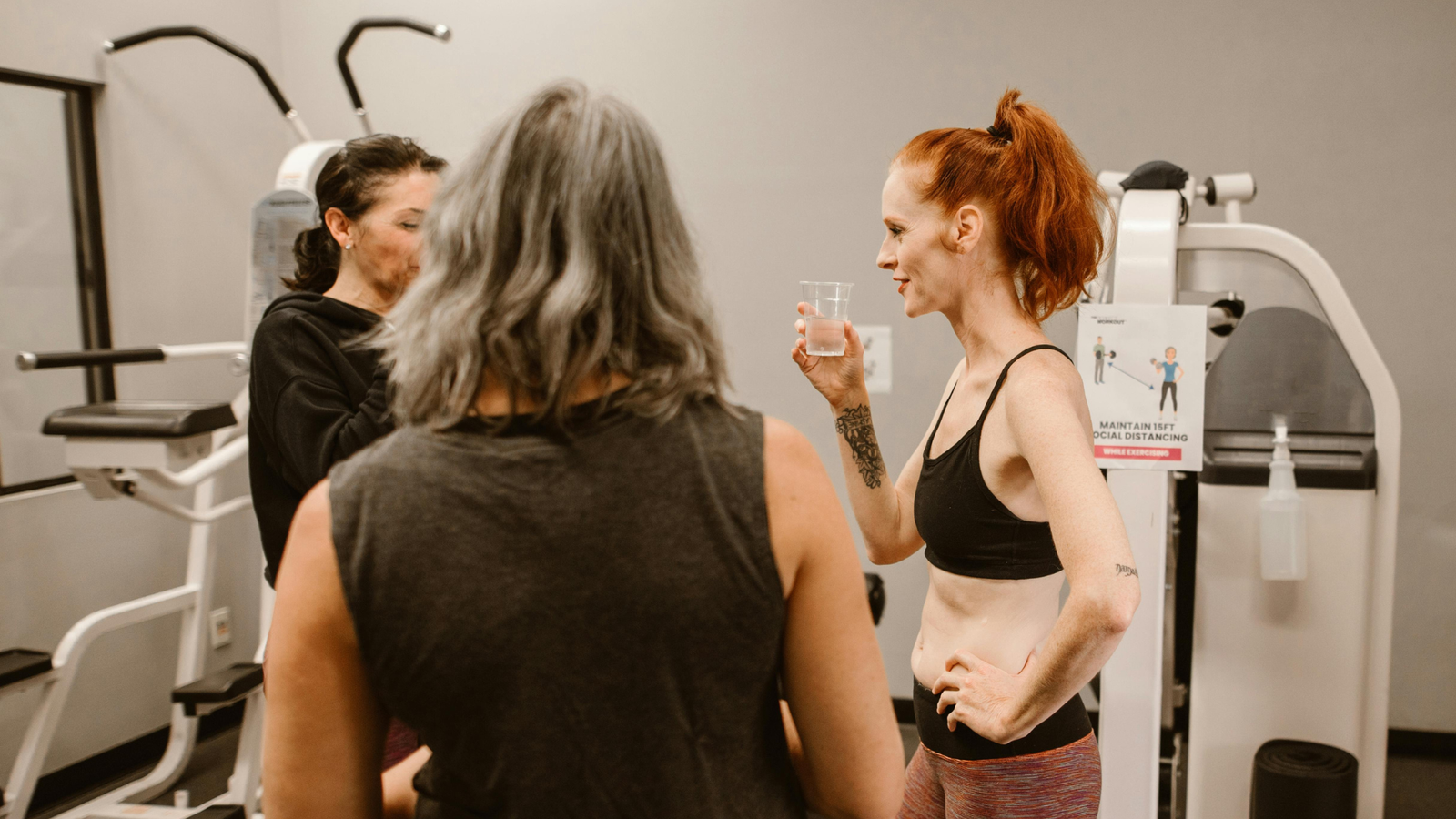 Three women chatting in a gym, one holding a water glass, with exercise equipment and a social distancing sign in the background.