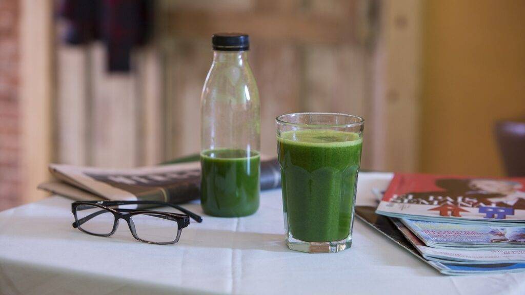 A glass and bottle filled with bright green smoothie or juice on a table, alongside reading glasses and magazines.